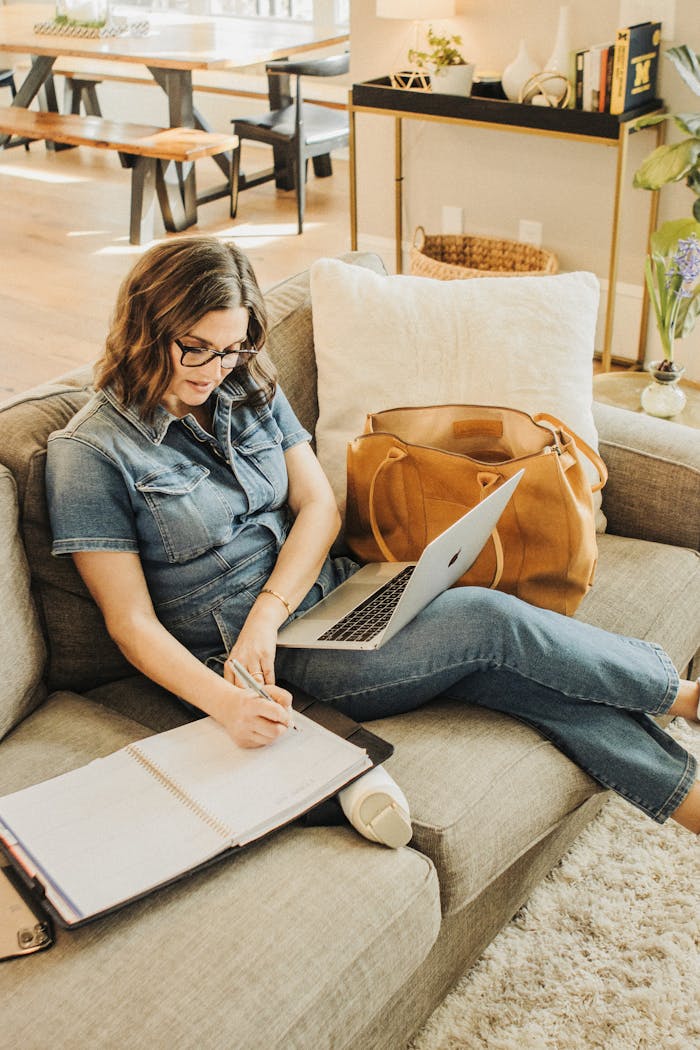 Woman in denim writing notes while working on a laptop with a leather tote bag beside her.