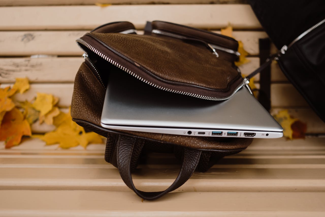 An open leather backpack with a laptop on a wooden bench surrounded by autumn leaves.